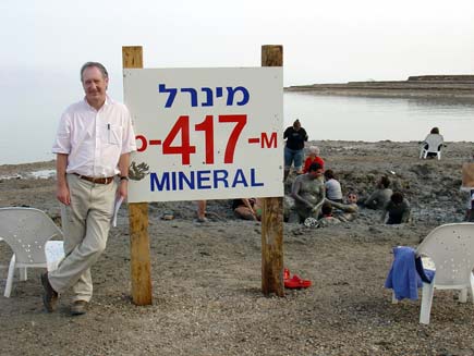 Roger Bolton standing in front of a notice with the Dead Sea in the background. Behind him, visitors bathe in a dip in the ground filled with mud