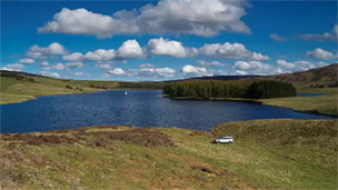 Colour view of a body of water set in a dip between rolling hills under a blue sky with a number of small white clouds. A group of trees sits on land projecting into the water and a silver car is parked in the foreground. .