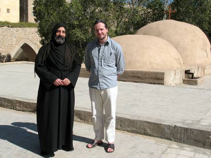 Father Iranaeus, dressed head to foot in black monastic robes with a scarf covering his head, with Nicholas Buxton in a stone courtyard inside the Monastery of Saint Macarius, with two domed roofs behind and below them