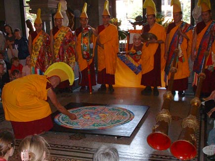 The monks are in full ceremonial dress now, wearing orange robes over their red tunics and orange and yellow crested headdresses. Tall Tibetan horn instruments are also visible. One monk has begun destroying the mandala by scraping his knuckle in a radial direction through the sand