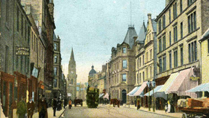 Hand-coloured view looking west along High Street, Perth. Various pedestrians can be seen in early 20th Century clothing. In the background are a double-decker tram and horse-drawn carts.