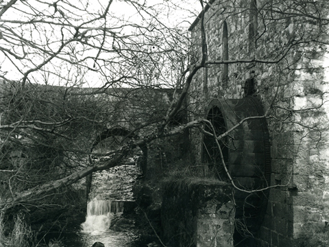 Black and white view of a watermill and wheel next to a narrow burn. An arched stone bridge crosses the burn over a small weir or waterfall.