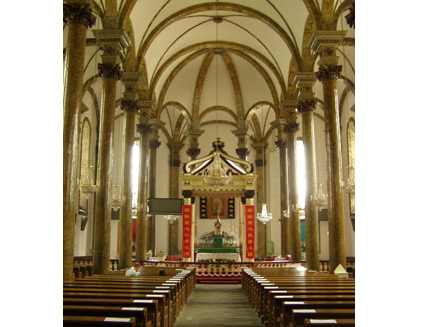Interior of the Eastern Cathedral, looking down the aisle of the east nave between rows of pews to an altar at the end. Beneath the high white ceiling and brown stone pillars, a single man is sitting praying