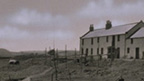 Black and white view of row of stone buildings with wheels, anchors and drying poles in the grass hollow below. A hill and bridges are seen in the in the distance.