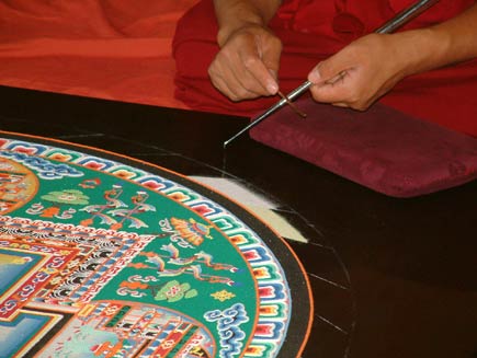 A closer view of the chak-pur rod. It is a metal tube about the length of the monk's forearm and the thickness of a pen, ridged along its side and tapered towards the end from which the sand is pouring. The monk is scraping a smaller rod along the chak-pur to make the sand flow smoothly