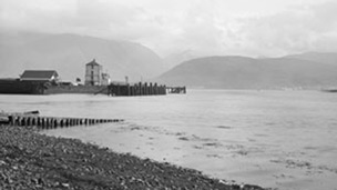 Black and white view from a rocky beach across Loch Linnhe to Ben Nevis. In the middle distance is a quayside featuring a tall, masonry constructed building and a wooden pier.