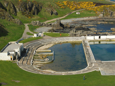 Colour view looking down from coastal cliffs to abandoned open air swimming complex. The pool to the left is partially dried up. The Art Deco pavillion to the left appears partially boarded up.