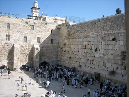 Old stone walls 15 metres tall, the remaining walls of the Second Temple. Jewish worshippers are gathered at the base of the huge wall, some wearing prayer shawls and standing up against the stone