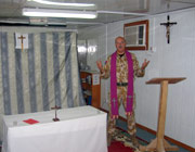 Chaplain Andrew Martlew in his chapel. The room is small and the furniture seems almost makeshift, with benches, a rug on the floor, a crucifix on the wall and a curtain behind the small altar to divide the room in two