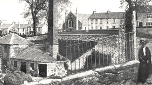 A woman in Edwardian dress stands next to a small set of mill buildings featuring a tall chimney stack. Behind is a row of dwellings and a church