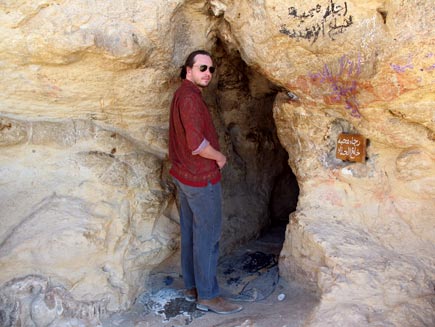 Nicholas Buxton at a small cave entrance cut in the striated yellow and beige stone. Writing in a non-Western script is visible next to the doorway