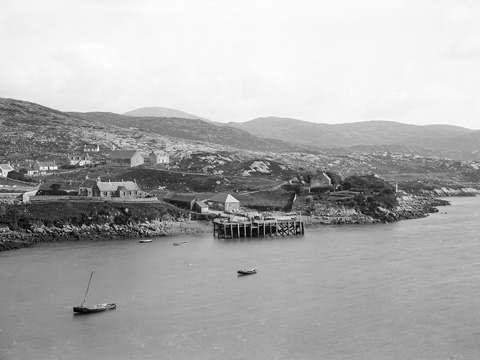 Black and white view across bay to a wooden pier with scattered cottages and other buildings behind.