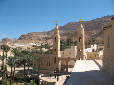 Saint Antony's monastery, a collection of warm-coloured stone buildings with small, arched windows. Two steeples topped by Christian crosses stand either side of the double arch entrance. Palm trees grow in small walled patches of green space outside the walls