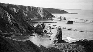 Black and white view looking down from cliffs to small harbour with buildings at Cove.