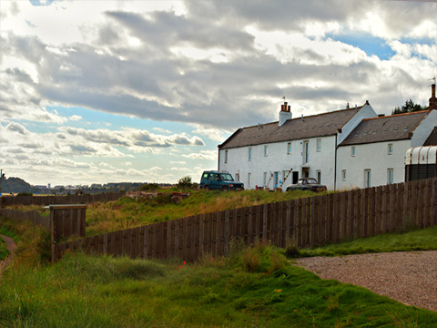 Coloure view of row of white-painted stone buildings with cars and a horse box parked in front. In the foreground is a modern wooden fence, which curves round a grassy area around the buildings. A footpath runs in to the distance along the left of frame.