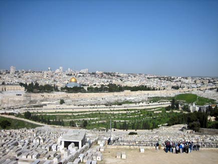 View of Jerusalem from high up, with the golden roof of the Dome of the Rock mosque clearly visible among the largely white buildings. In the foreground a group of people is looking at the view from a hilltop Jewish cemetary