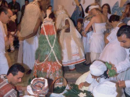 Crowd of people in a room. Foreground shows people on the floor preparing food in leaves. Main background shows man, dressed in white, standing up with rafia fringe covering his face, dancing.