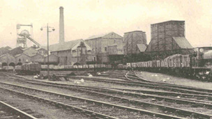 A steam train with a long line of wagons sits on one of a number of railway lines in front of industrial buildings including a winding tower and large chimney stack.