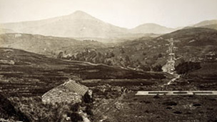 Black and white view showing a small stone shelter and a flat concrete area with mountains behind.
