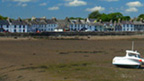 Small boats sit beached in a harbour where the tide is out. A row of single and two-storey buildings line the promenade behind.