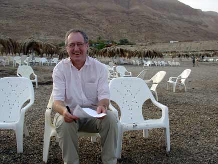 Roger Bolton sits on a plastic chair on the beach, holding a script