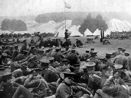 Black-and-white photo from 1915. Soldiers in First World War uniform sit on the ground, some looking at the camera, while a Church of England clergyman speaks. In the background are rows of tents