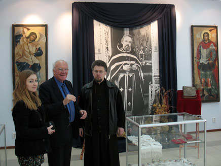 Maria Grigorenko, Michael Bordeaux and Victor Grigorenko in the Father Alexander museum, standing among glass cases in front of a huge photograph of Alexander in full Orthodox vestment