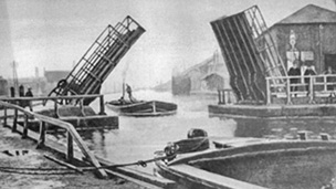 Black and white view from canal side showing a barge passing under an open bascule bridge.