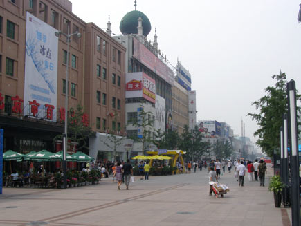 Wangfujing shopping precinct, a wide pedestrian area surrounded by tall buildings. Shoppers and outdoors café tables are on the pavement