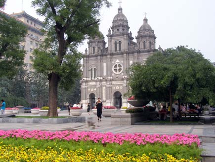 The Eastern Cathedral, a white stone building topped with three small domed towers. Tubs and beds of flowers decorate the square in front of the building while people sit at tables in the shade of a tree