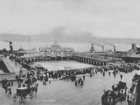 Black and white view of Dunoon Pier lined with crowds of people. Horses and carriages are on the street in the foreground. Two steamers with smoking chimneys are moored in the background.