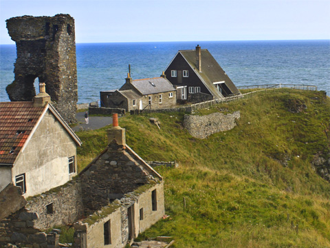 Colour view of a rocky peninsula on which stands the ruin of Old Slains Castle. Beyond this are a renovated traditional cottage and a 20th Century A-Frame house. In the foreground are the roofless ruin of another traditional cottage with a 20th century pitch-roofed cottage beside it.