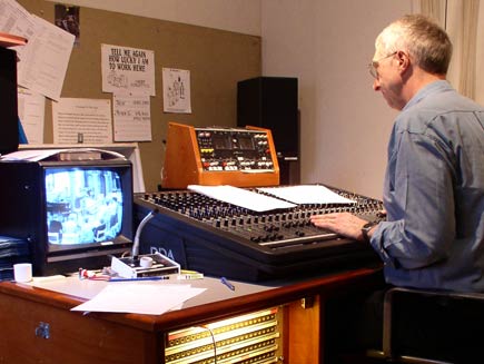 Looking over the shoulder of the sound engineer, who is balancing sound levels using the console in front of him. A monitor on the desk shows the scene in the main room of the church. On the far wall, a noticeboard displays a few clippings and cartoons, including one with the caption 'Tell me again how lucky I am to work here'