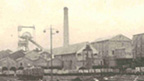 A steam train with a long line of wagons sits on one of a number of railway lines in front of industrial buildings including a winding tower and large chimney stack.