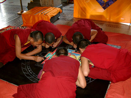 A wider shot shows five monks crowded around the mandala-in-progress, which is about two metres in diameter. They are kneeling on cushions placed around the tabletop, which is close to ground level