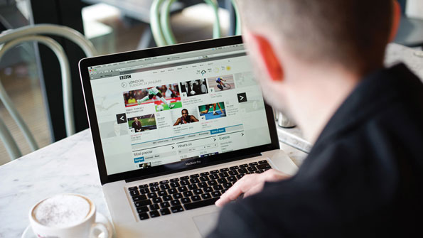 Looking at the homepage on a white laptop in a cafe, over the shoulder of a blurred patron. There is a cappucino on the mock marble tabletop. 