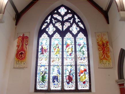 A tall stained glass window designed by William Morris, depicting eight saints. On either side of the window are hangings showing a person with the flame of the Holy Spirit, captioned 'Prayer'