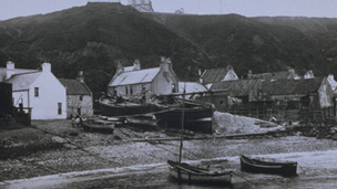 Black and white view showing small and large fishing boats pulled up onto the shore with cottages beneath tall cliffs behind.