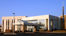 Modern, square, light-coloured building with a triangular-roofed porch at the front and a large Christian cross displayed on a pillar