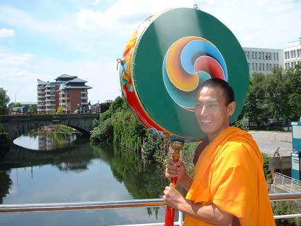 A smiling monk crosses the River Irwell with a large, brightly-painted instrument that looks somewhat like a tambourine or drum, somewhat like a giant lollipop