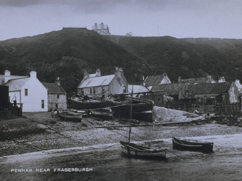 Black and white view showing small and large fishing boats pulled up onto the shore with cottages beneath tall cliffs behind.