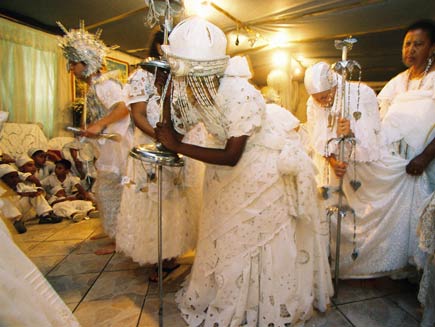 Three women stand bent, dressed in white, clutching metal walking sticks.