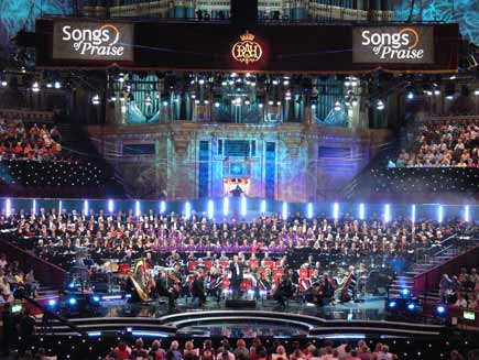 Choirs and orchestra on stage, with the organist visible in an illuminated alcove above the choirs. The stage, designed on sweeping curved lines, is illuminated with coloured lights in varying patterns