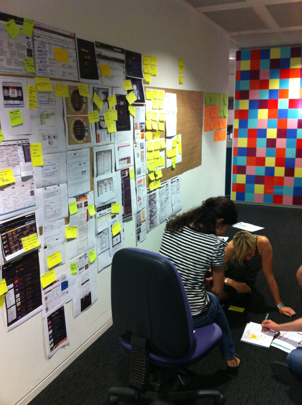 A meeting wall full of ideas and potential designs. Two people lean over a third writing in a book.