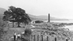 Black and white view of a couple with a pram and dog walking along a winding, coastal footpath. In the background are trees and the tall, cylindrical Battle of Largs Monument.