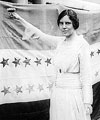 Alice Paul in 1920, standing in front of suffrage flag holding up a glass