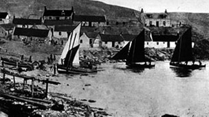 Black and white view of small sailing boats landing on a beach. A cluster of cottages rises up the hill behind.
