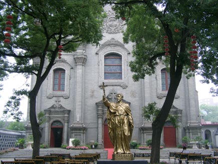 The white stone Southern Cathedral is visible behind a gold-coloured statue of a figure holding a cross, which stands in the middle of an outdoor seating area. Red Chinese lanterns hang from the trees that shade the seats