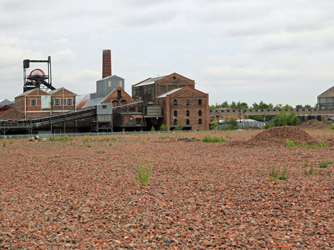 Industrial buildings including a winding tower and large chimney stack with an open area of red gravel in the foreground.