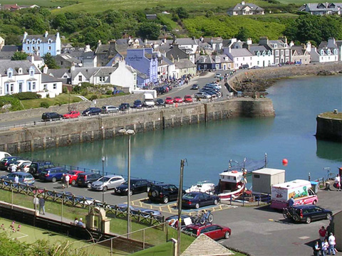 Colour view of harbour containing three boats. Cars and an ice-cream van are parked around the harbour walls. The edge of tennis courts can be seen in the foreground.